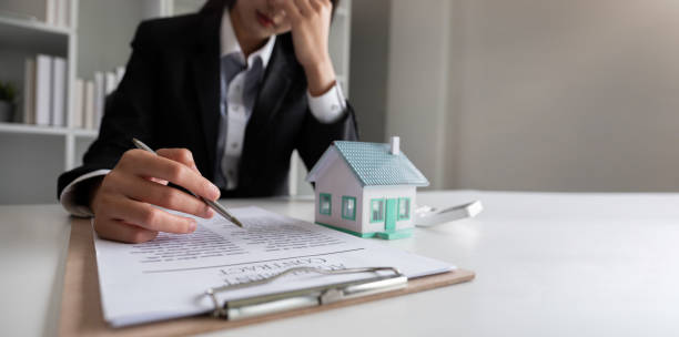 A frustrated businessperson reviews a contract at a desk with a small model house on it.